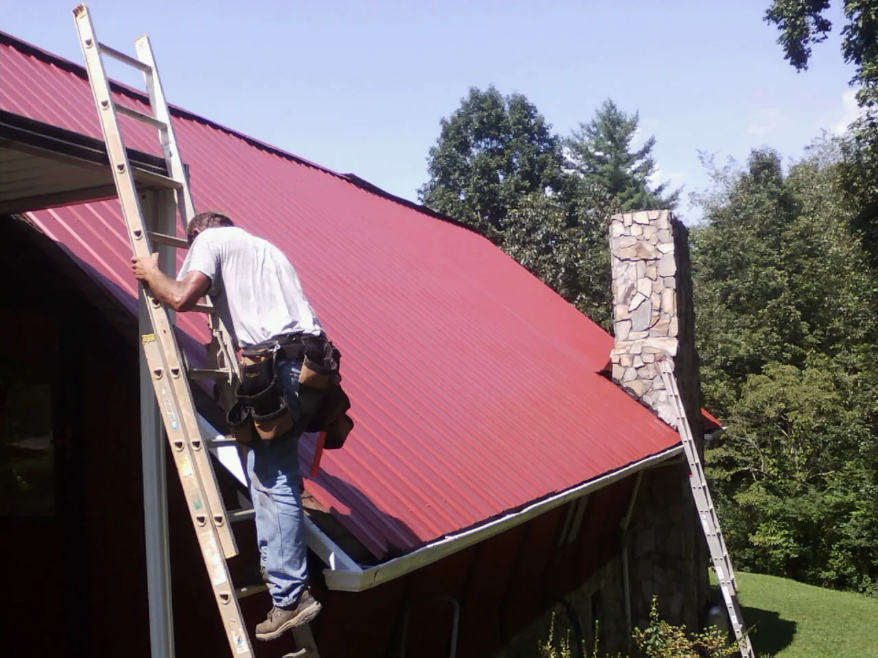 Roofer on a ladder beside a red metal roof and stone chimney — Soffit & Fascia Repair by Roofing Brothers in Doty
